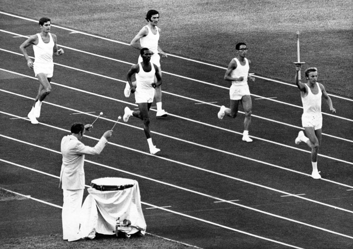 Schwarz-Weiß-Fotografie einer Gruppe von Läufern, der vorderste trägt die olympisch Fackel