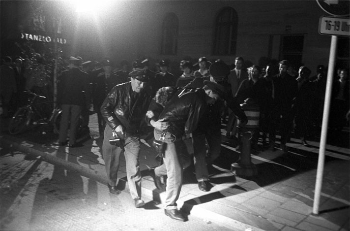 Schwarz-Weiß-Fotografie von drei Polizisten die einen Mann abführen, vor einer Menschenmenge