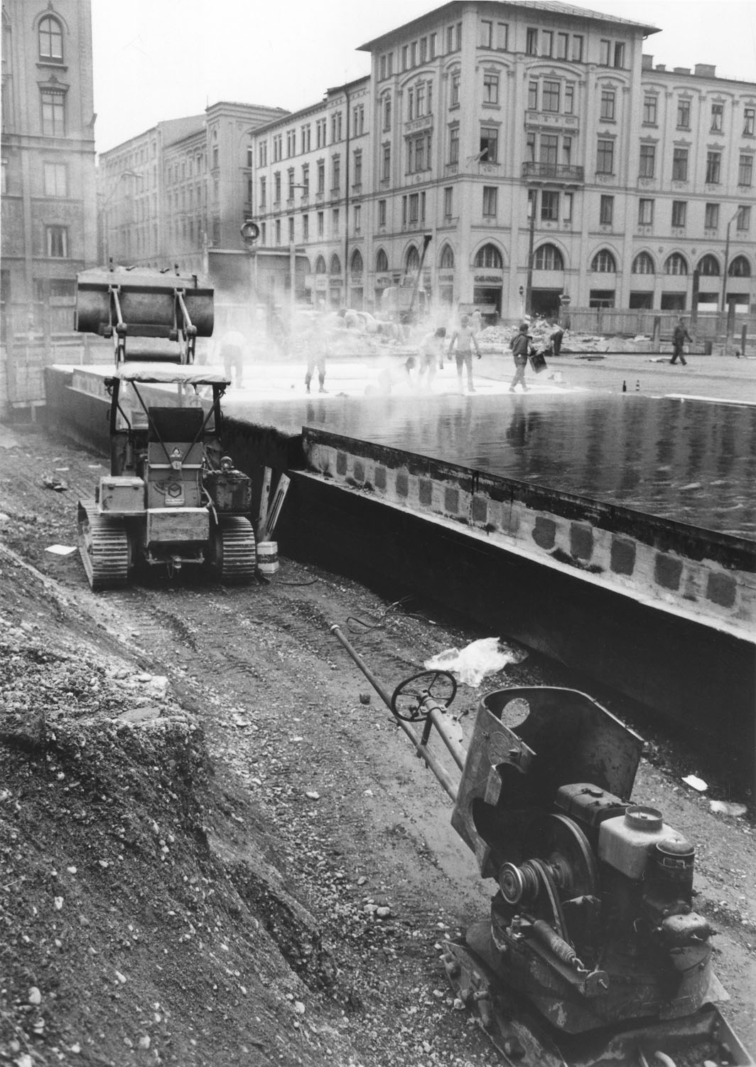 Schwarz-Weiß-Fotografie einer Baugrube mit einer Baumaschine. Im Hintergrund wird Wasser versprüht.