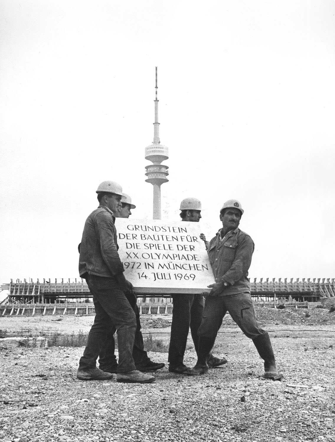 Schwarz-Weiß-Fotografie einer Bauarbeitergruppe die ein schweren Betonstein durch eine Baustelle tragen. Im Hintergrund steht ein Fernsehturm.