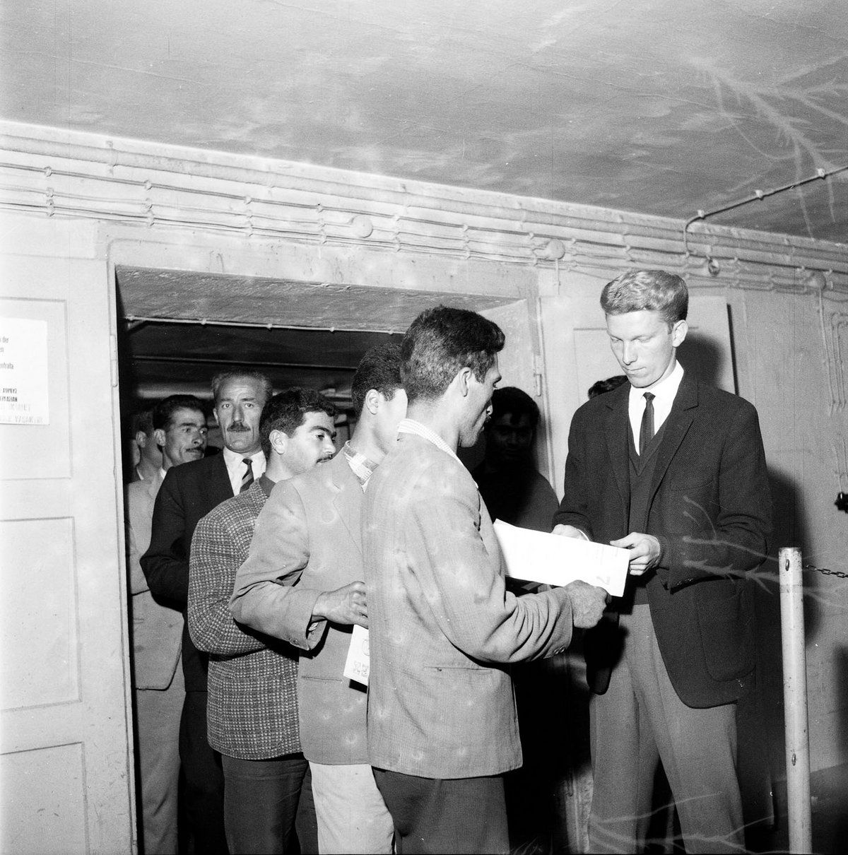Schwarz-Weiß-Fotografie einer Gruppe Männern, die anstehen und Papiere in der Hand halten.