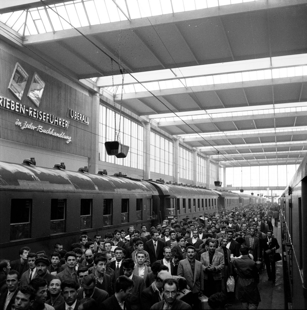 Schwarz-Weiß-Fotografie eines Bahnhofs mit einer drängelnden Menschenmasse. Zudem stehen leere Züge an den Gleisen.