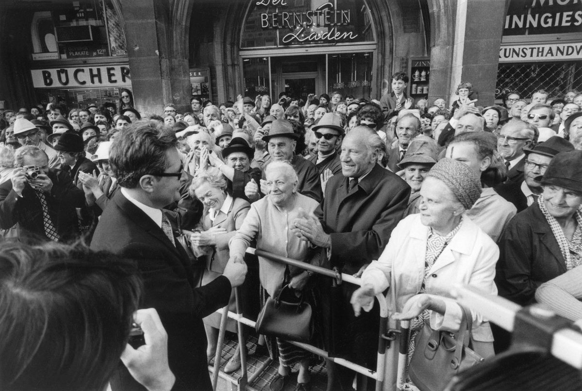 Schwarz-Weiß-Fotografie einer großen Menschenmenge. Im Vordergrund schüttelt ein Mann einer älteren Dame die Hand.