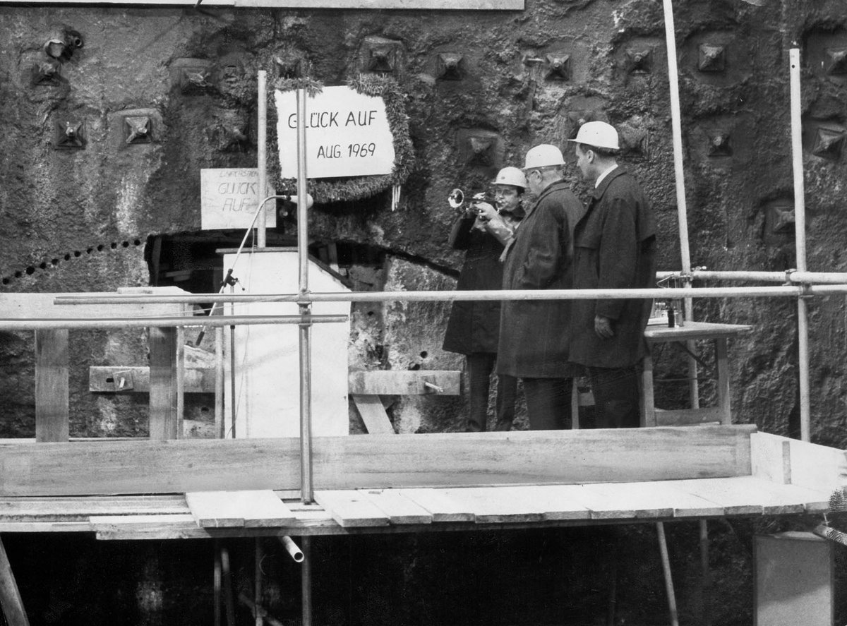 Schwarz-Weiß-Fotografie von drei Männern auf einer Baustelle. Im Hintergrund steht ein Schild mit “Glück auf Aug 1969“.