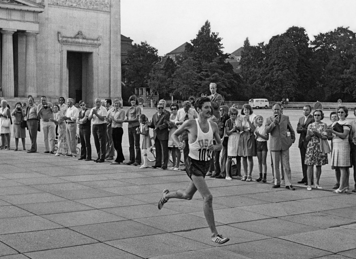 Schwarz-Weiß-Fotografie eines laufenden Mannes, im Hintergrund stehen jubelnde Menschen.