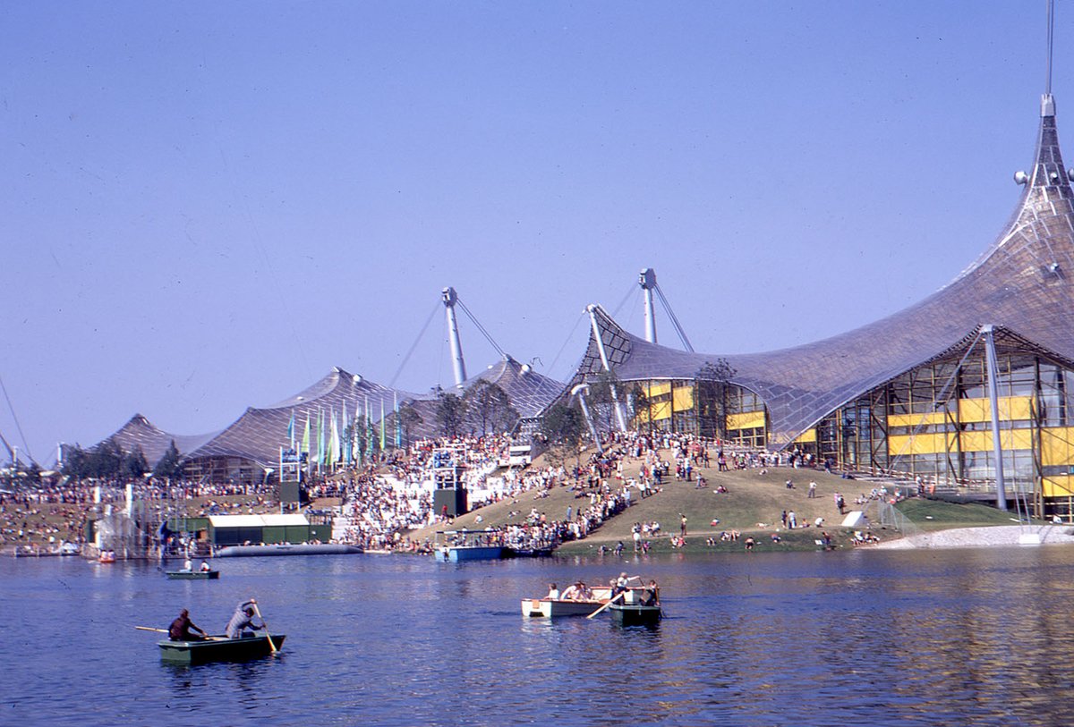 Fotografie von einem See und einem Stadion, in der Mitte befinden sich viele Menschen sitzend auf einem Berg