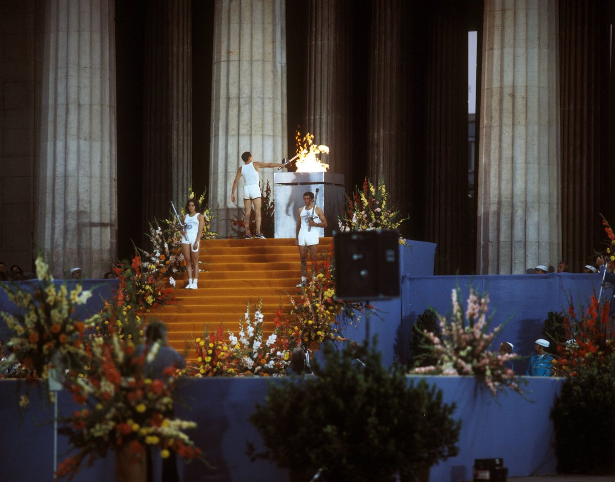 Fotografie einer Tribüne mit Blumen dekoriert, im Hintergrund entzündet ein Mann eine Flamme.