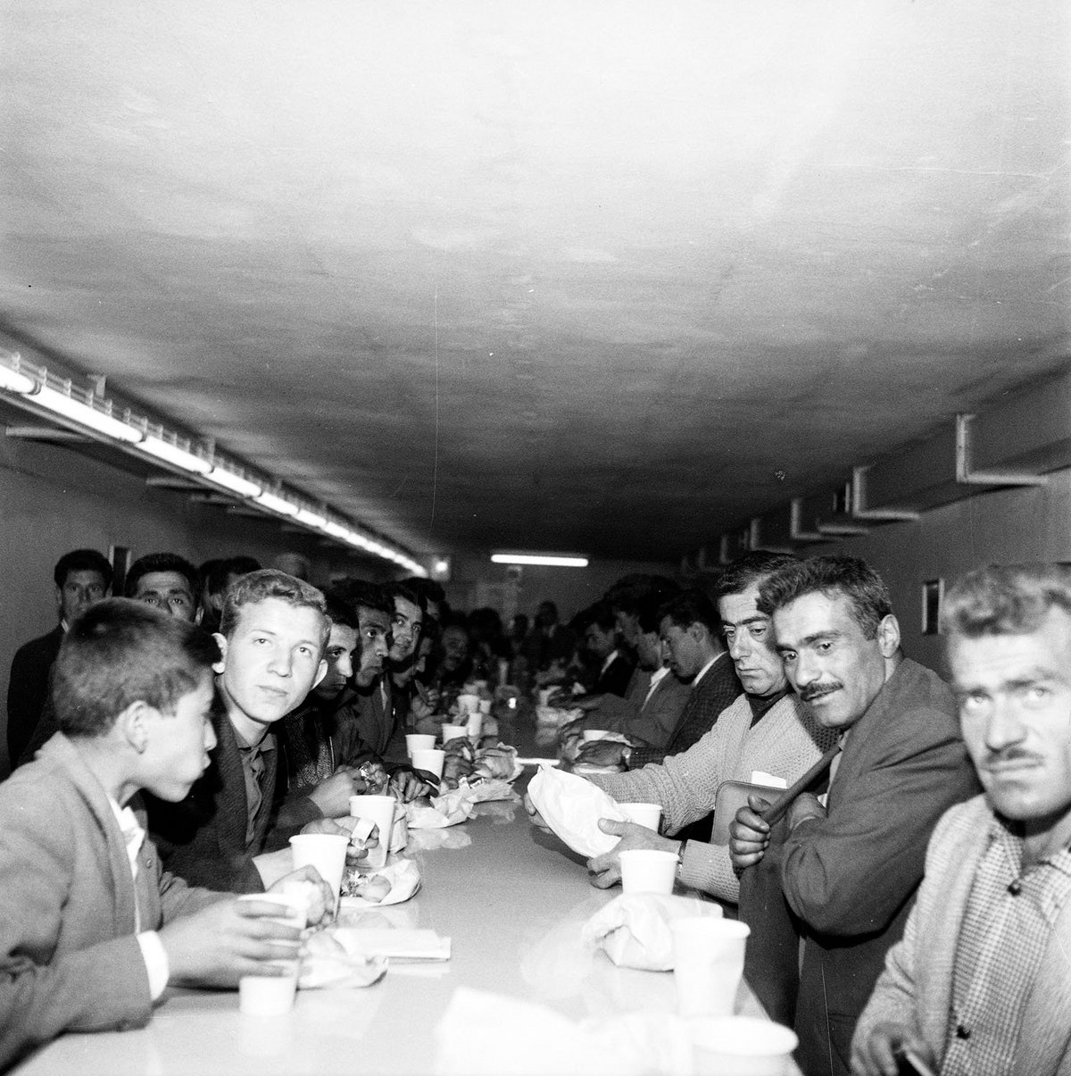 Schwarz-Weiß-Fotografie einer Gruppe Männern, die an einem Tisch essen und trinken.