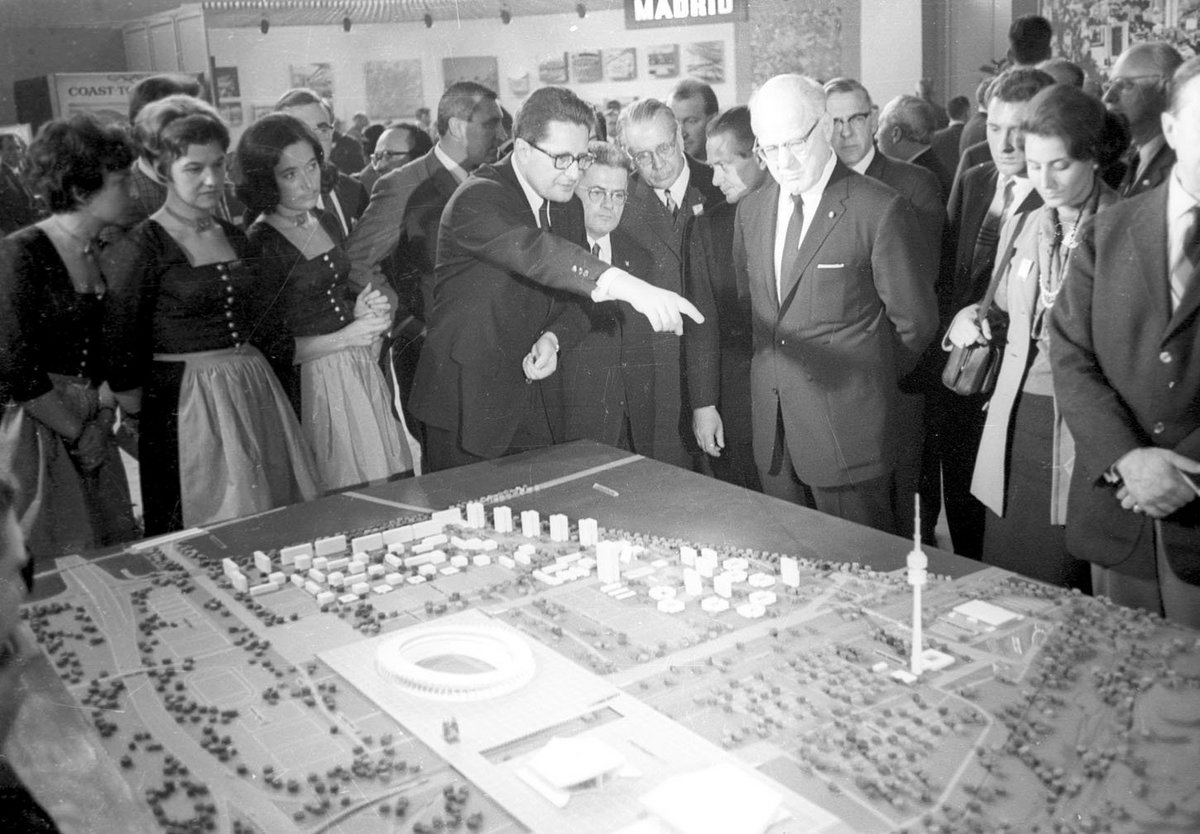 Schwarz-Weiß-Fotografie einer Gruppe Menschen. Ein Mann deutet auf ein Modell des Olympiapark München