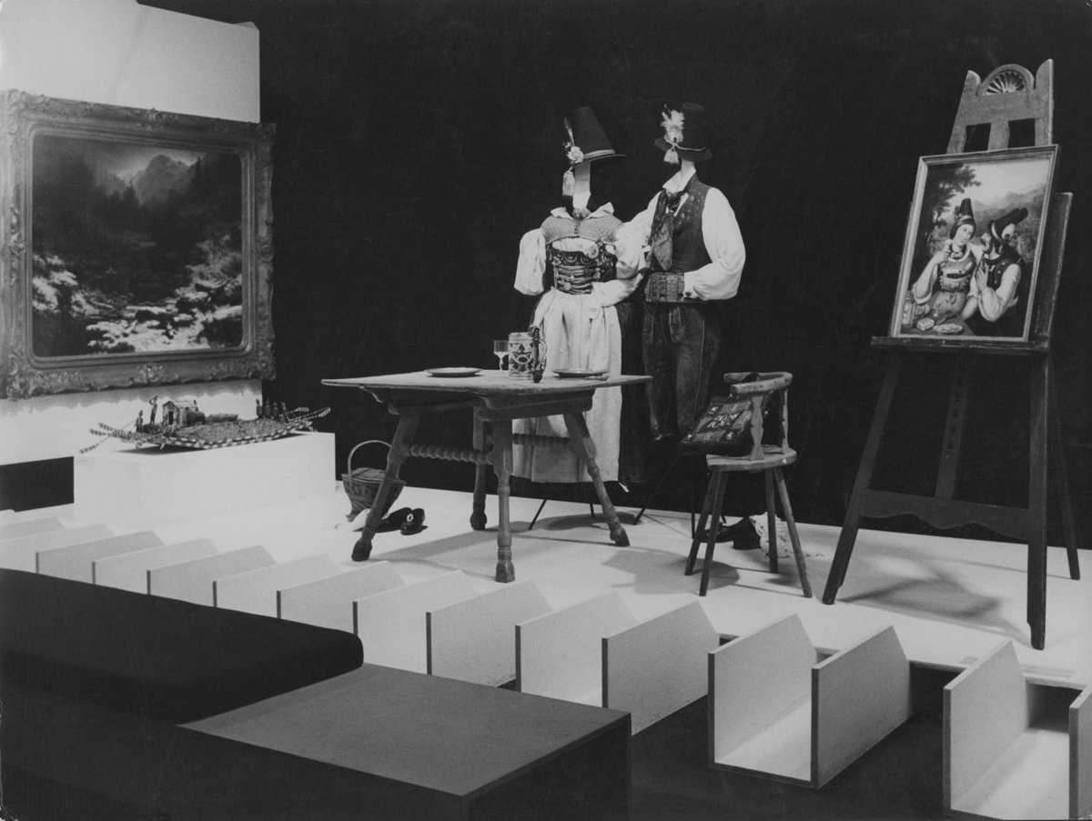 Schwarz-Weiß-Fotografie einer Ausstellungsansicht mit bayerischen Trachten, Gemälden und Möbeln. 