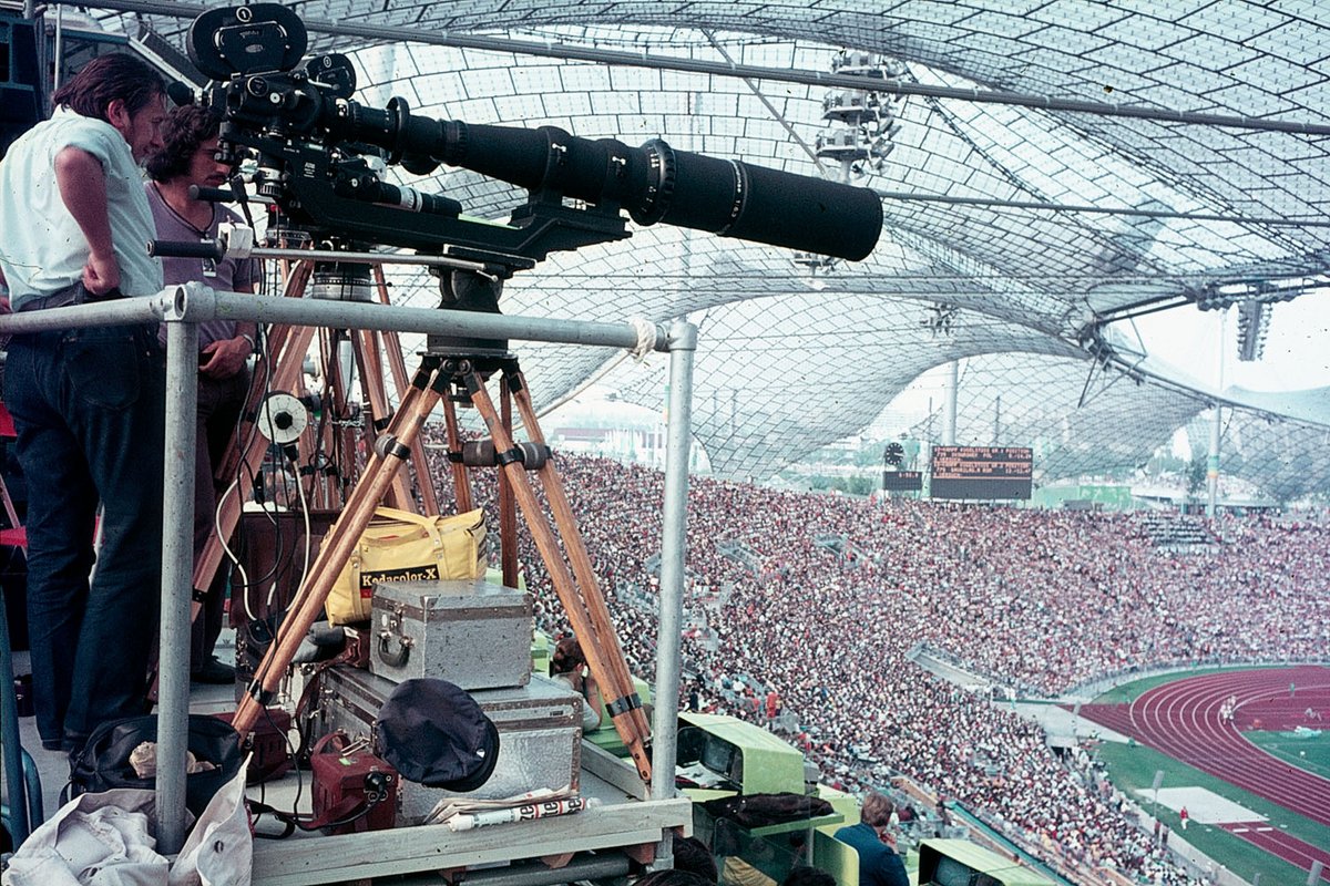 Fotografie eines vollbesetzen Stadion von Innen. Im Vordergrund stehen zwei Männer mit einer Filmkamera. 